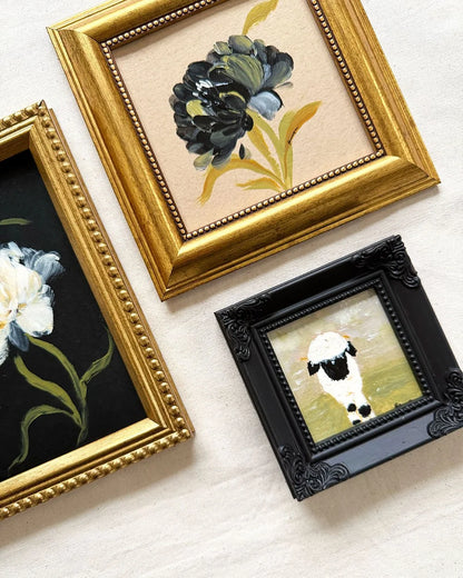 Three framed art original arranged on a light beige textured wall. The top original features a dark blue and green flower in a gold frame.