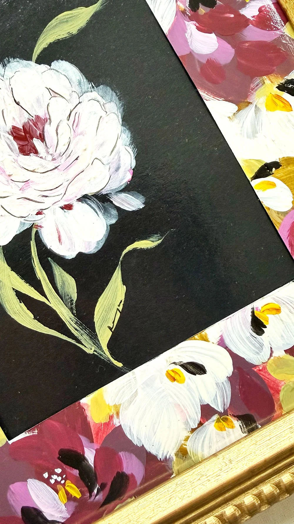 A close-up of a framed floral artwork showing a large white flower with green leaves on a black background, with additional multicolored floral sections and a gold frame visible.