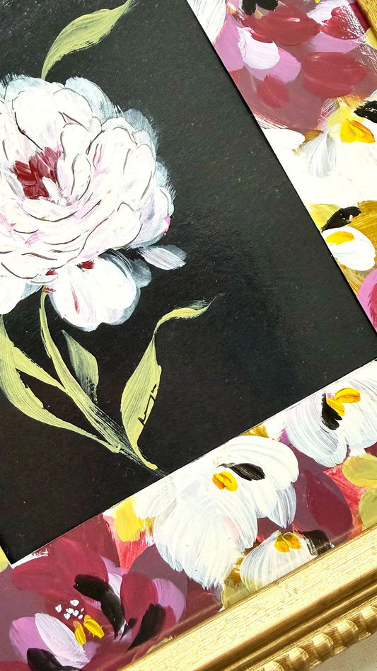 A close-up of a framed floral artwork showing a large white flower with green leaves on a black background, with additional multicolored floral sections and a gold frame visible.
