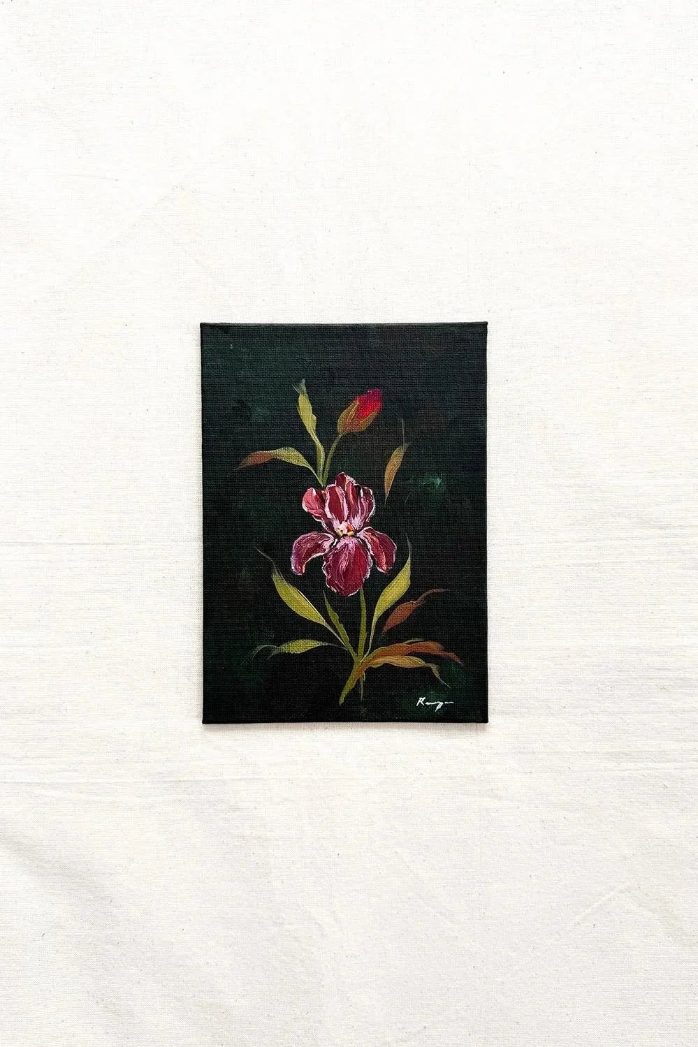 A small original framed painting of a red and pink iris flower with green and yellow leaves on a dark background, displayed on a textured white fabric surface.