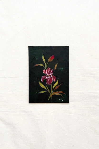 A small original framed painting of a red and pink iris flower with green and yellow leaves on a dark background, displayed on a textured white fabric surface.