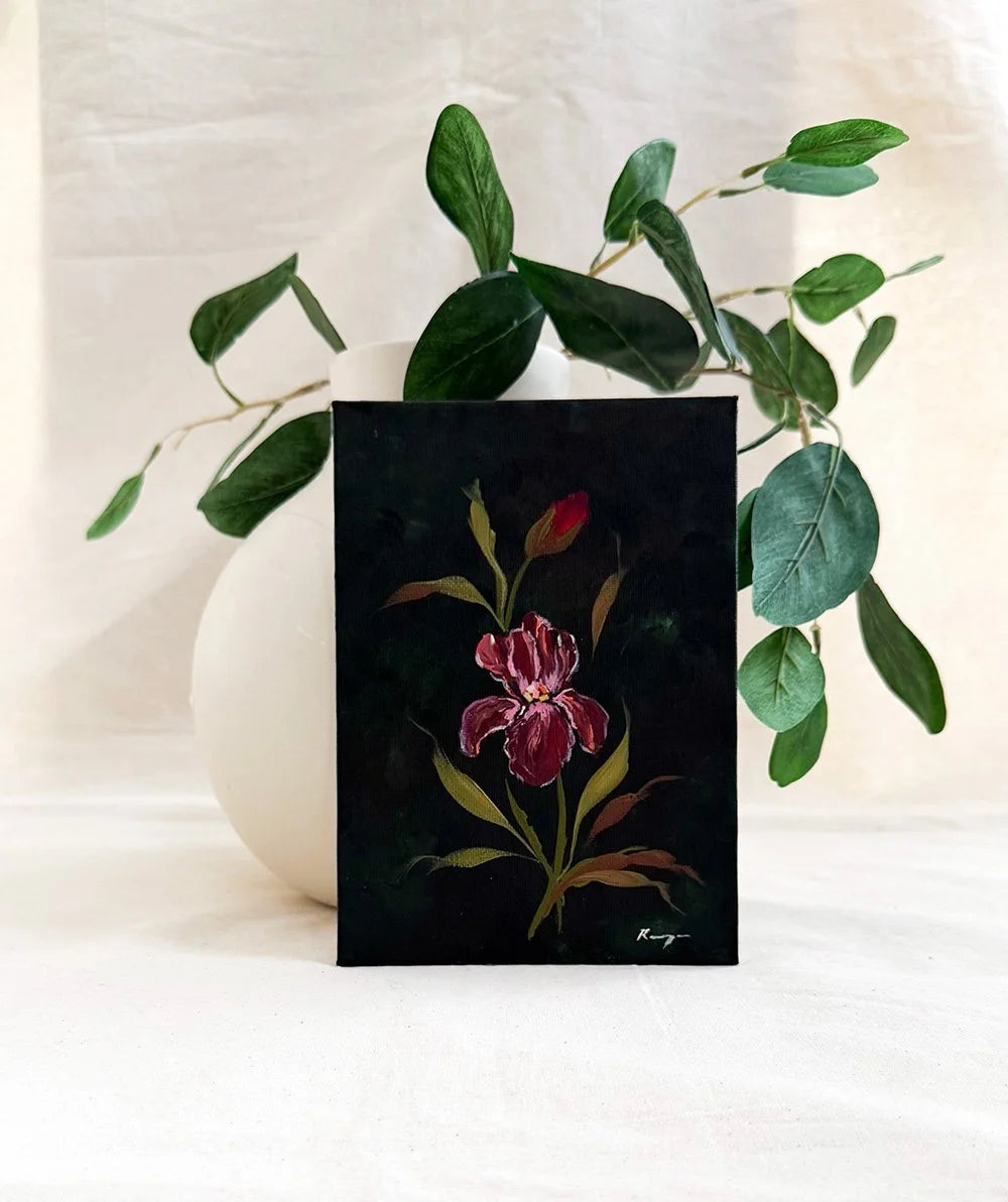 A dark green art print featuring a painted red iris flower with green and gold leaves, styled in front of a white ceramic vase with eucalyptus branches against a draped white fabric background.