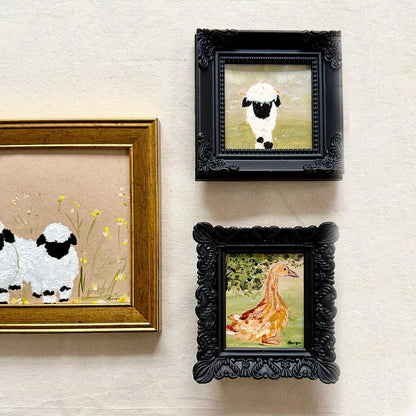 Three framed animal paintings hanging on a textured beige wall. The left frame has a gold border and shows two white sheep with black faces in a field of yellow flowers.