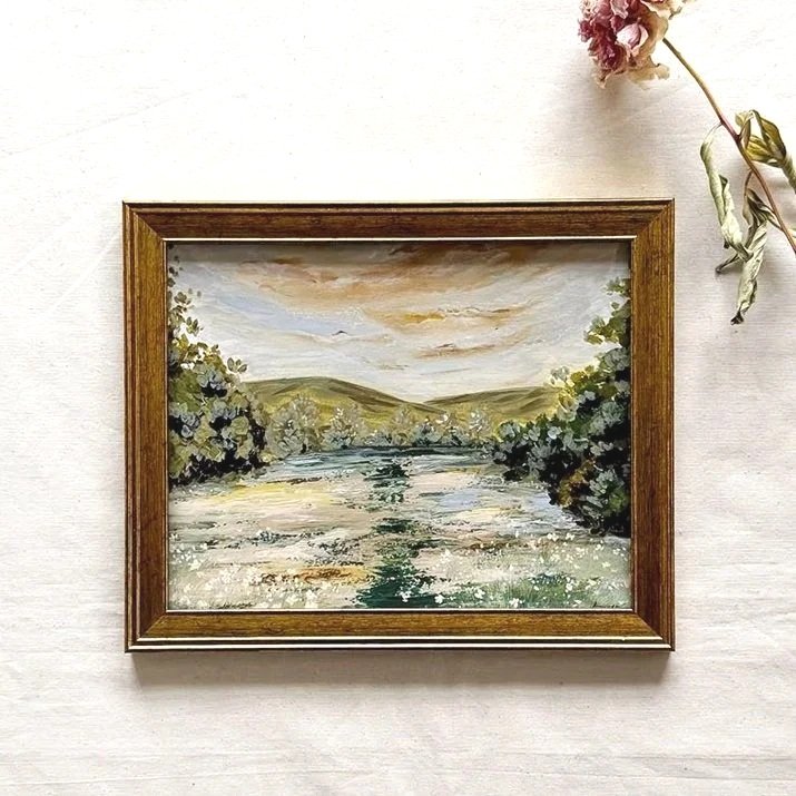 A framed original landscape painting by Ramya Rajiv featuring a grass flowing through a valley with trees and rolling hills under a cloudy sky, housed in a rustic brown wooden frame 