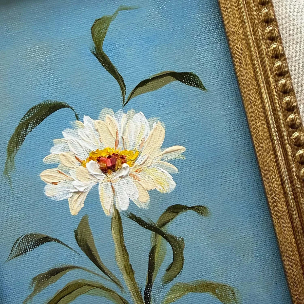Close-up of a framed painting featuring a white daisy with a yellow and red center on a light blue textured background, with green leaves and stems
