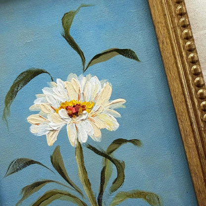 Close-up of a framed painting featuring a white daisy with a yellow and red center on a light blue textured background, with green leaves and stems