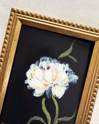 Framed floral painting featuring a white flower with green leaves on a black background, enclosed in a decorative gold frame.