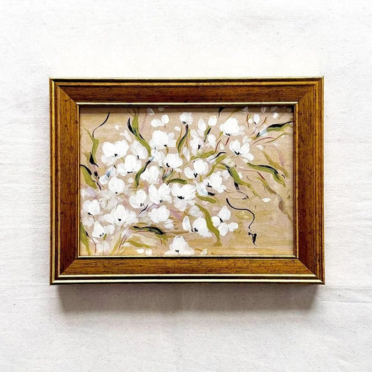A framed painting of white flowers with green stems on a beige background, housed in a warm brown wooden frame 