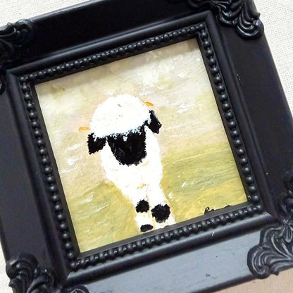 A framed original painting of a black and white sheep on a textured cream and yellow background, set in an ornate black frame.