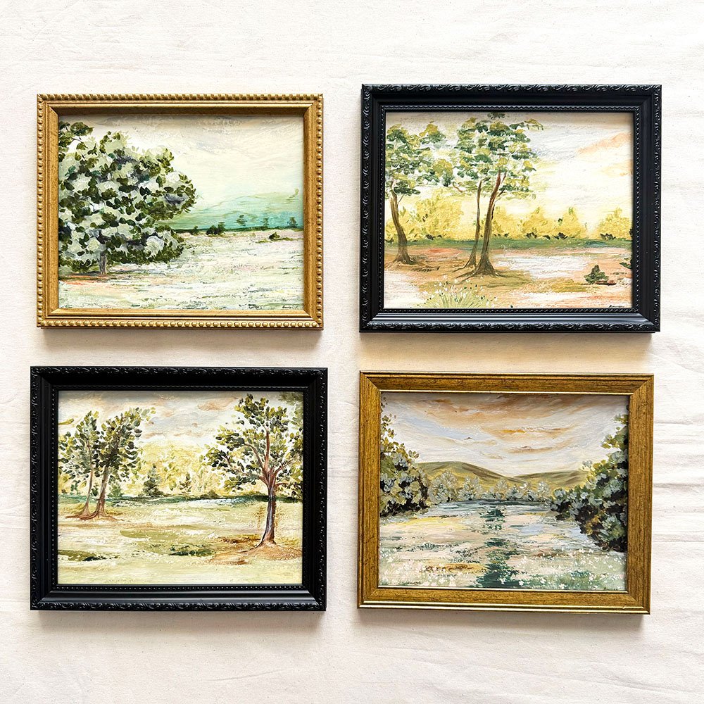 Four framed original landscape paintings arranged in a 2x2 grid on a textured off-white wall. Each painting depicts a serene outdoor scene with trees, fields, and distant hills, and they are housed in different frame styles: gold, black, and two mixed wood finishes.