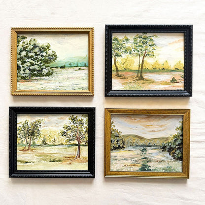 Four framed original landscape paintings arranged in a 2x2 grid on a textured off-white wall. Each painting depicts a serene outdoor scene with trees, fields, and distant hills, and they are housed in different frame styles: gold, black, and two mixed wood finishes.