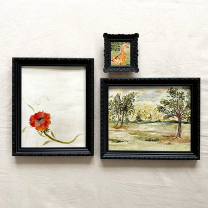 Three framed original wall art pieces a large black frame with a red flower on white paper, a smaller black frame with a giraffe painting, and a medium black frame with a landscape painting of trees.