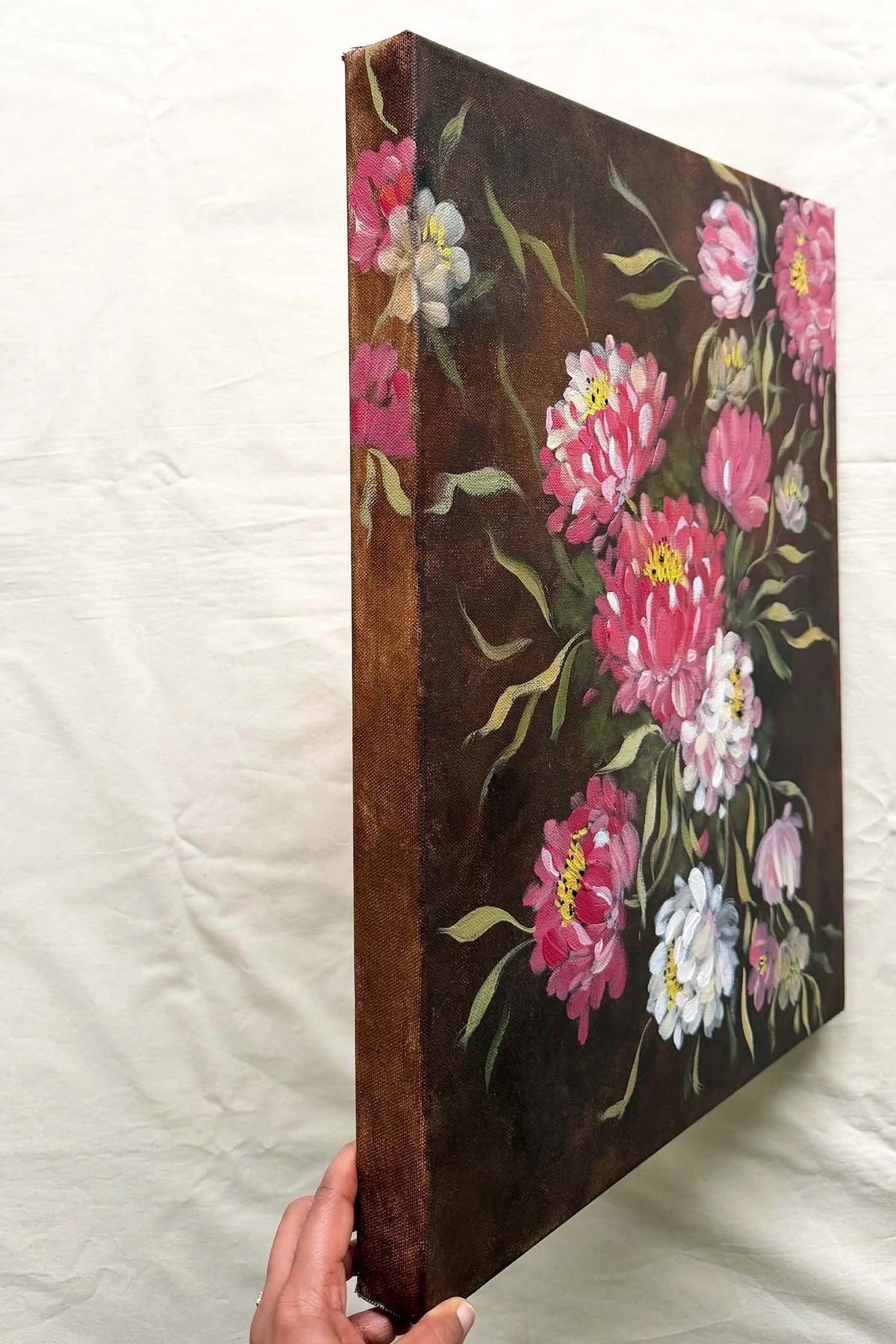 A gallery wrapped canvas painting showing a floral design with pink, white, and yellow flowers on a dark brown background. 