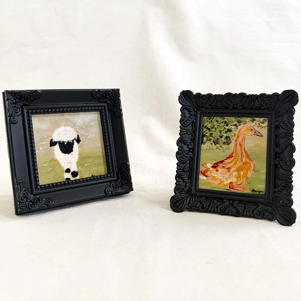 Two small framed paintings on a white fabric background. The left frame shows a black-and-white sheep, and the right frame shows a yellow-orange duck with green foliage.