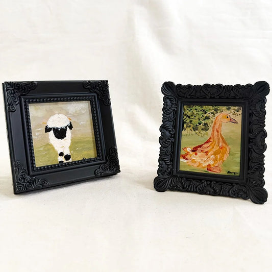 Two small framed paintings on a white fabric background. The left frame shows a black-and-white sheep, and the right frame shows a yellow-orange duck with green foliage.