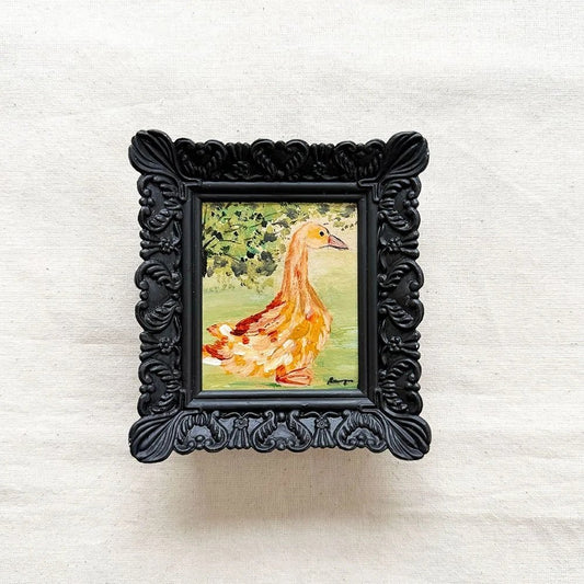A small framed painting of a duck. The artwork features a yellow-orange duck with green foliage behind it, set within an ornate black frame.