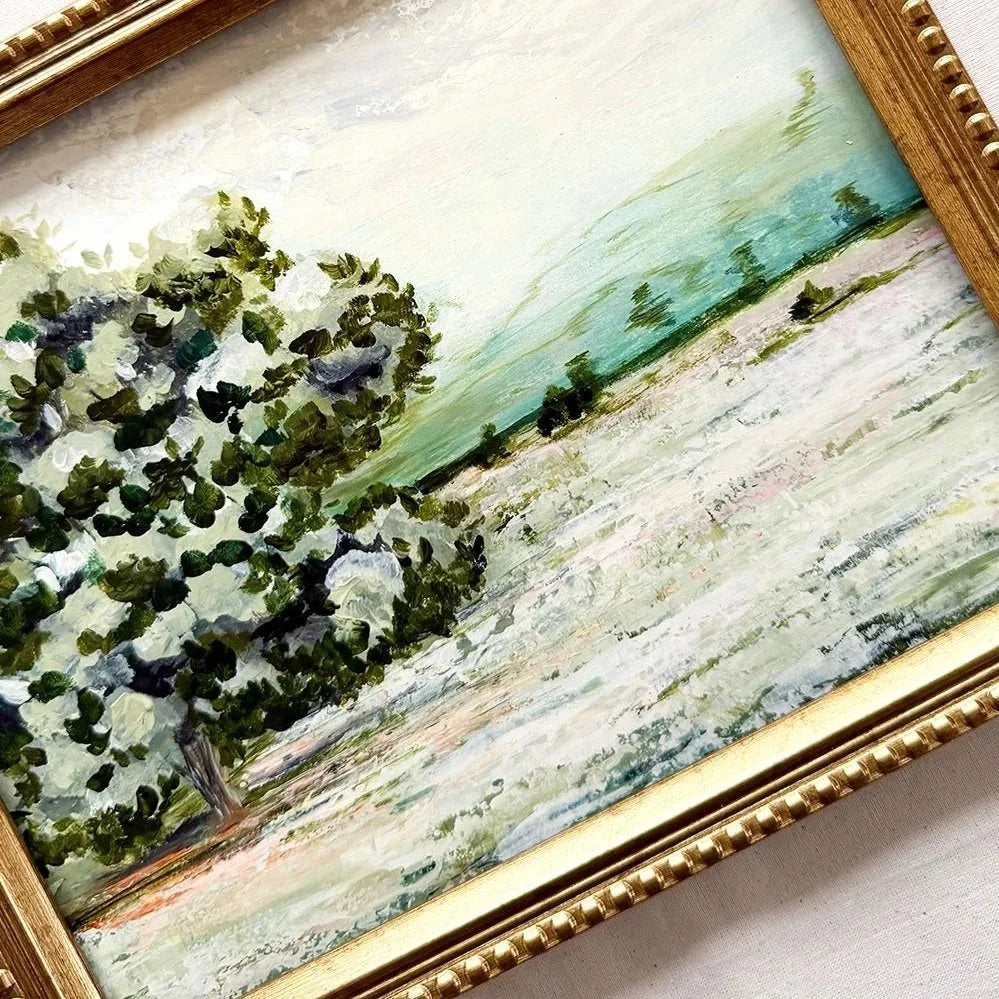 A framed landscape painting featuring a green tree on the left and a soft green and white field under a pale sky, enclosed in a gold-toned ornate frame.