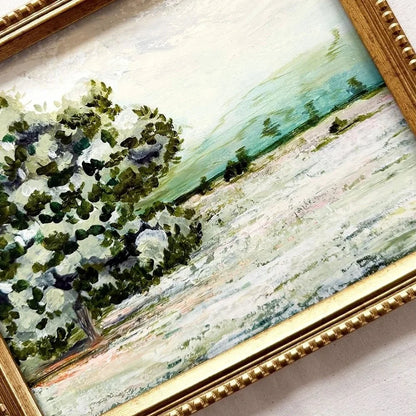 A framed landscape painting featuring a green tree on the left and a soft green and white field under a pale sky, enclosed in a gold-toned ornate frame.