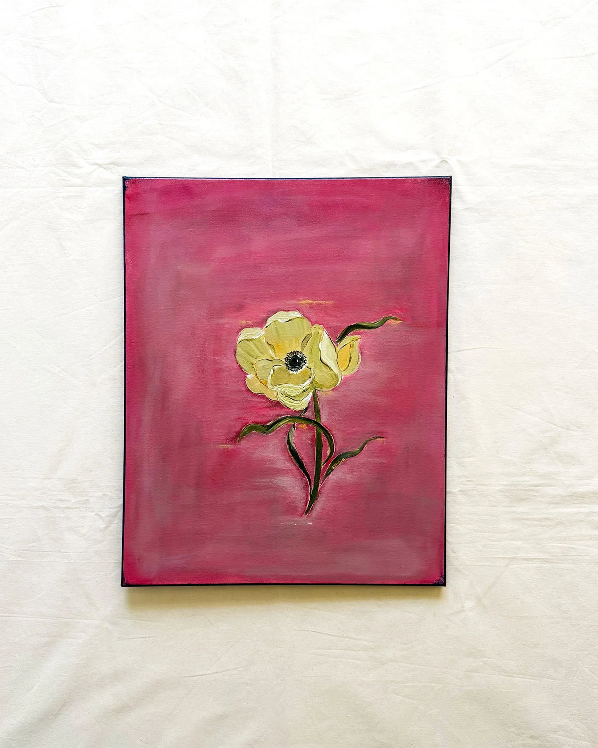 A square painting of a single pale yellow flower with dark green stems on a textured magenta-pink background, displayed against a wrinkled off-white fabric backdrop.