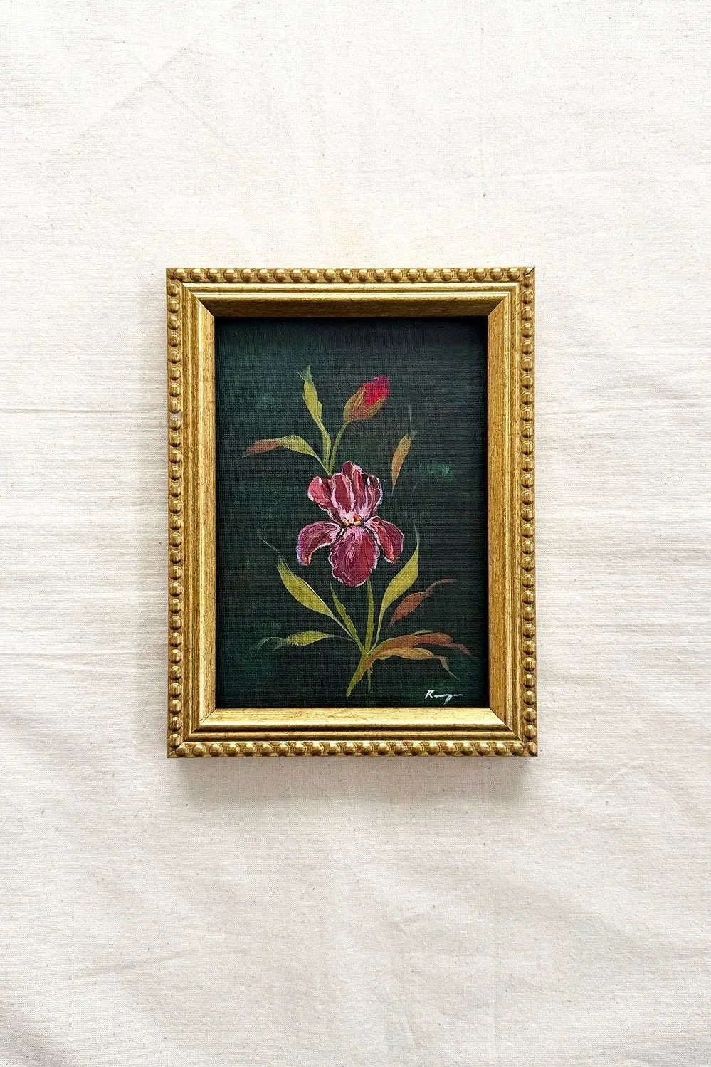 A small framed original of a pink iris flower with green and yellow leaves on a dark background, set in a decorative gold frame 