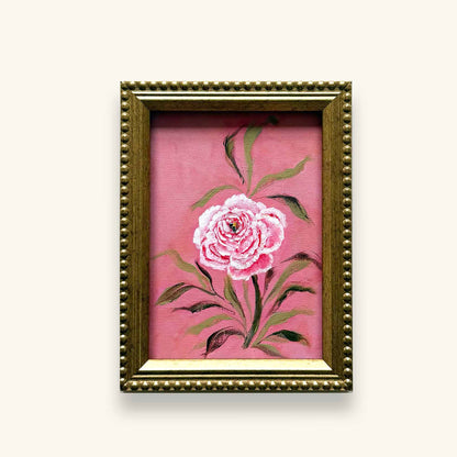 A framed original June flower of the month art featuring a pink rose with green leaves on a pink background, enclosed in an ornate gold frame 
