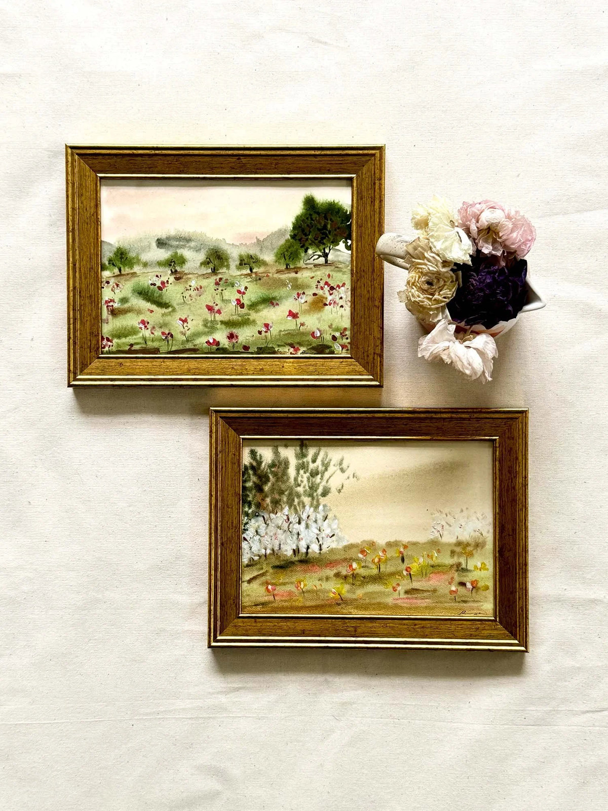 Two small framed landscape paintings with gold frames hanging on a light fabric background, alongside a small bouquet of pink, white, and dark purple flowers.