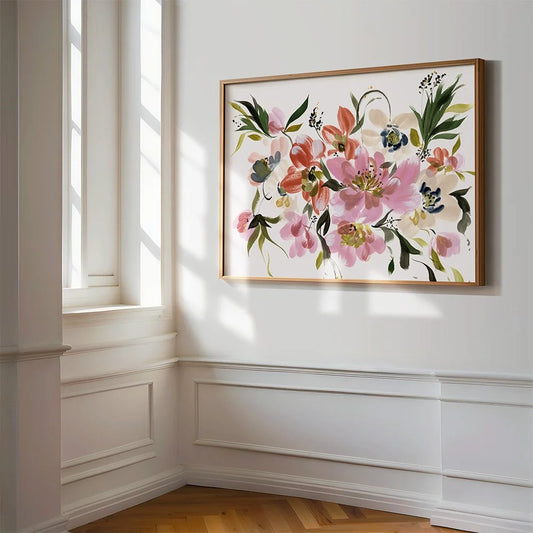 A framed floral art print hangs on a white paneled wall in a bright room with a window and wood floor. The artwork features pink, peach, green, and cream flowers with blue accents inside a thin gold-toned frame.
