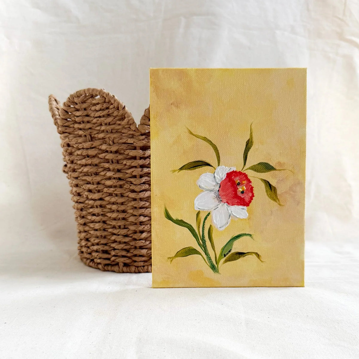 A original floral art painting with a yellow background, featuring a white Daffodil with a red center and green leaves, displayed next to a woven brown basket 