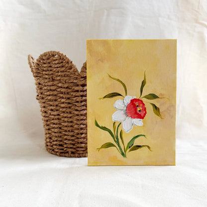 A original floral art painting with a yellow background, featuring a white Daffodil with a red center and green leaves, displayed next to a woven brown basket 