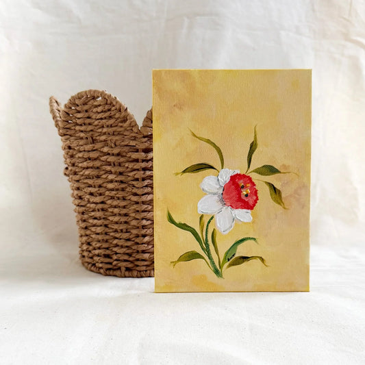 A original floral art painting with a yellow background, featuring a white Daffodil with a red center and green leaves, displayed next to a woven brown basket 
