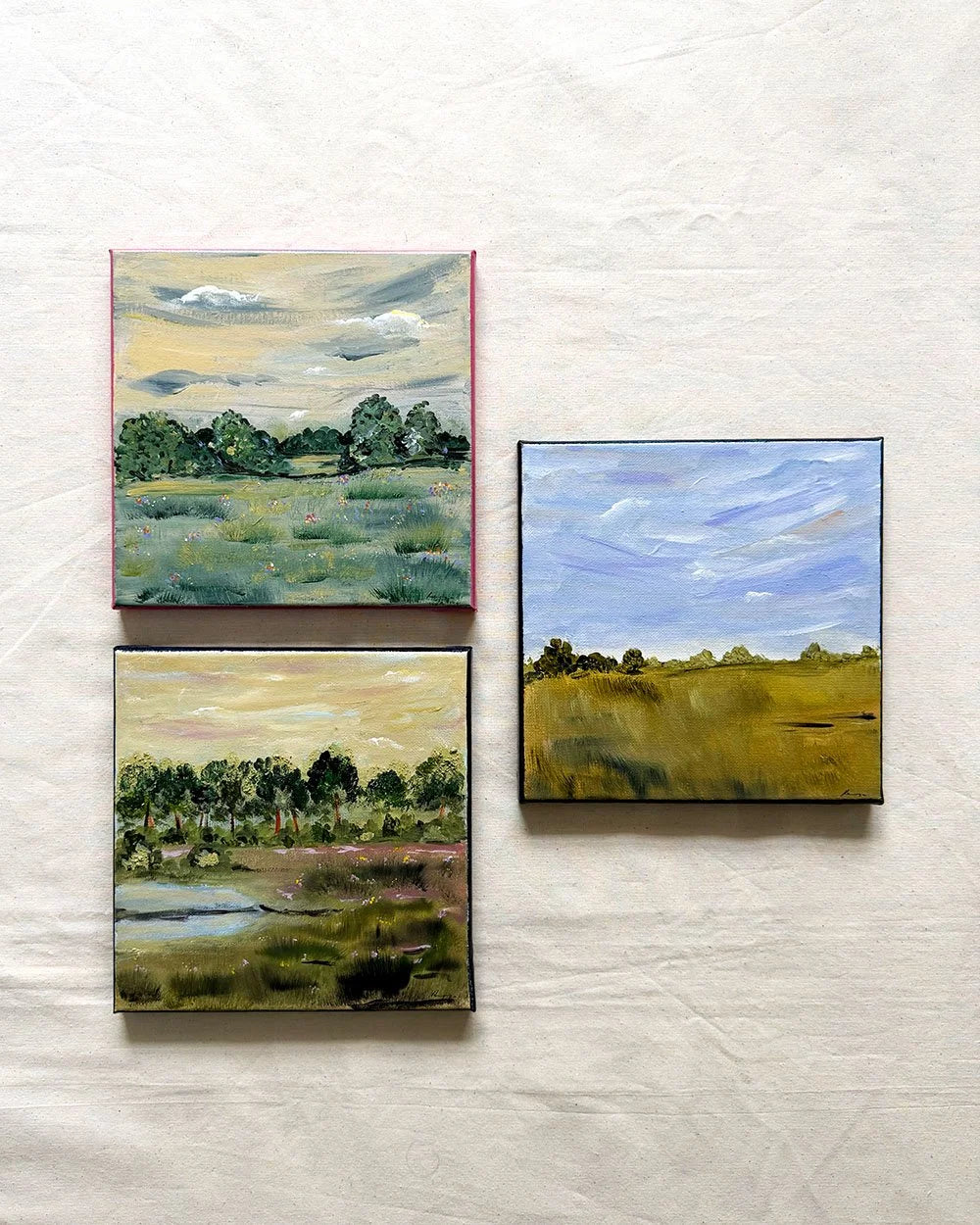 Three small landscape paintings displayed on a textured off-white fabric background. Two paintings on the left show green fields and trees under a pale sky, and one painting on the right shows a blue sky over a golden field.