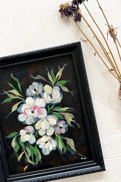 A framed floral painting featuring white and pink blossoms with green leaves on a dark background, set in a black ornate frame on a light fabric surface with dried flowers as props.