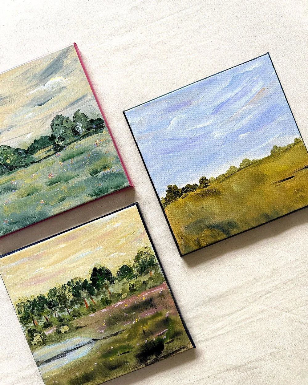 Three small landscape paintings arranged on a light fabric background. The paintings depict grassy fields, trees, and sky scenes in muted green, blue, yellow, and pink tones.