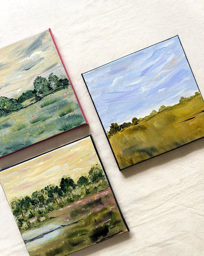 Three small landscape paintings arranged on a light fabric background. The paintings depict grassy fields, trees, and sky scenes in muted green, blue, yellow, and pink tones.