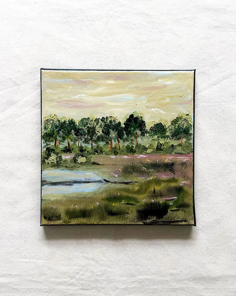 A square landscape painting with a black frame, depicting a grassy field, trees, and a river under a pale sky, mounted on a textured white wall.