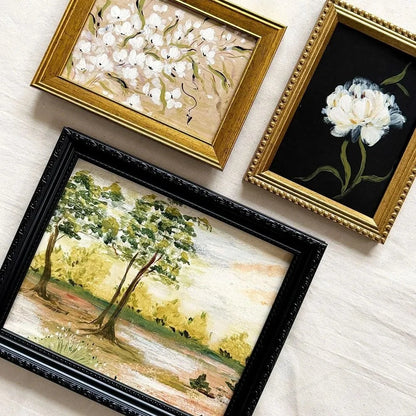 Three framed paintings arranged on a light fabric background: a black-framed landscape with trees, a gold-framed white floral painting, and a gold-framed black floral painting with a white flower.