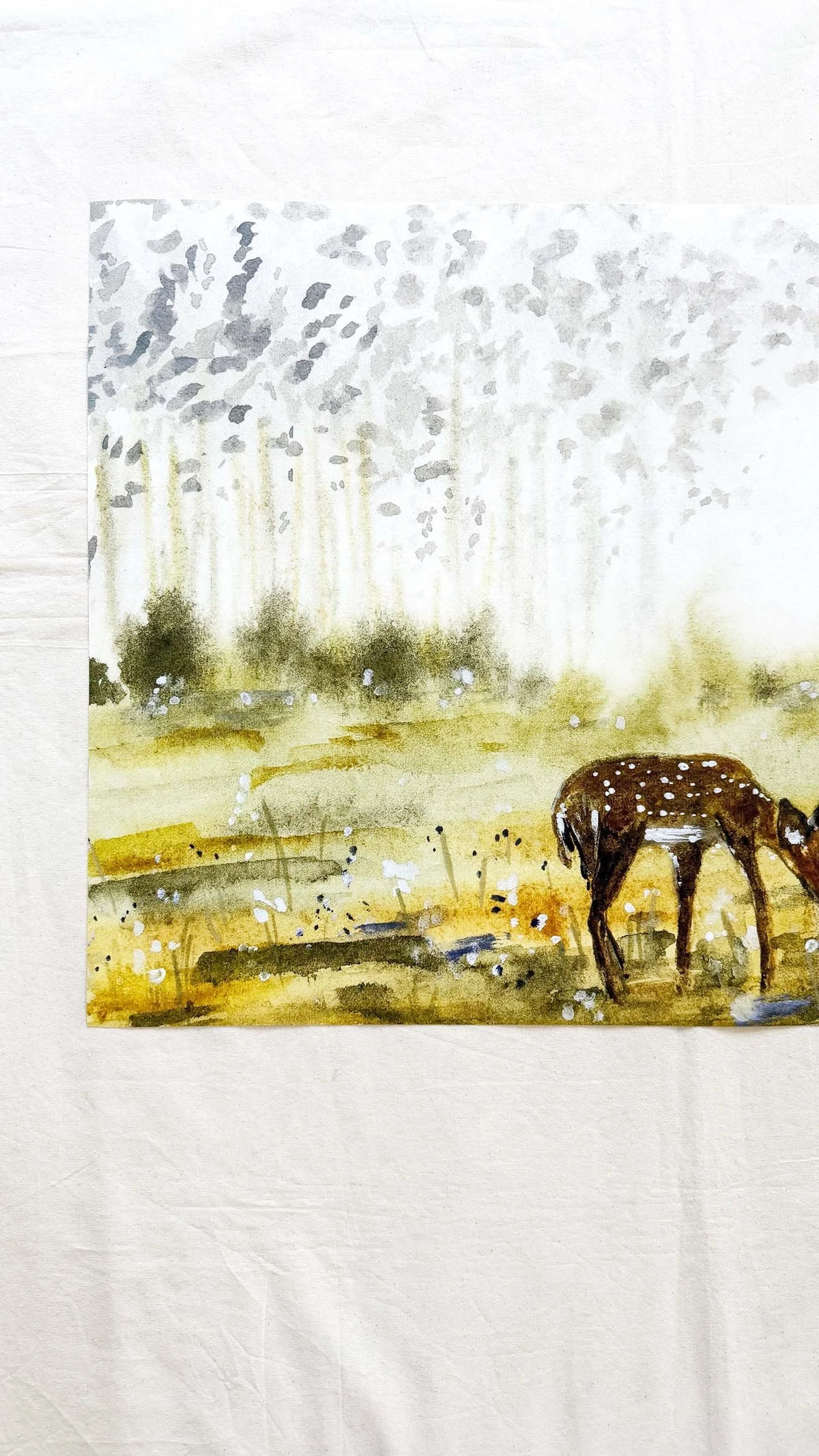 A square watercolor painting of a spotted deer in a grassy field under a misty forest, displayed on a textured white fabric background.