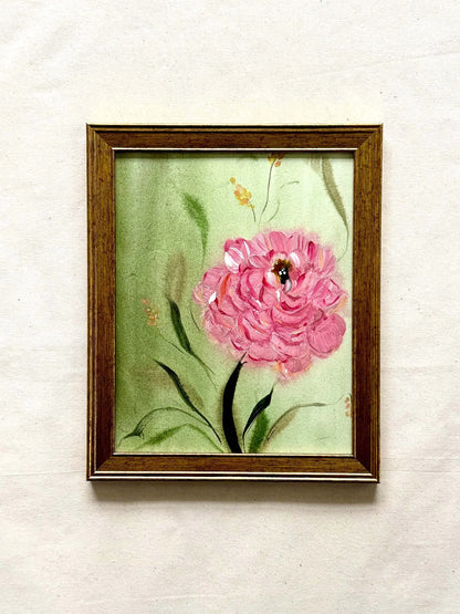 A framed painting of a pink flower with green leaves on a light green background, enclosed in a brown wooden frame and mounted on a textured off-white wall.