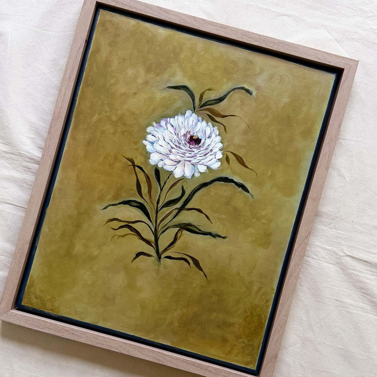 Framed original floral artwork featuring a white flower with dark green leaves on a textured olive-gold background, set in a light wood frame.Art by Ramya Rajiv