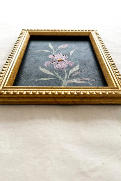 A framed floral original featuring a pink iris with green leaves on a dark gray background, enclosed in an ornate gold frame 