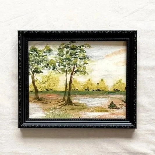 A framed landscape painting featuring three trees by a river, with a black ornate frame original by Ramya Rajiv