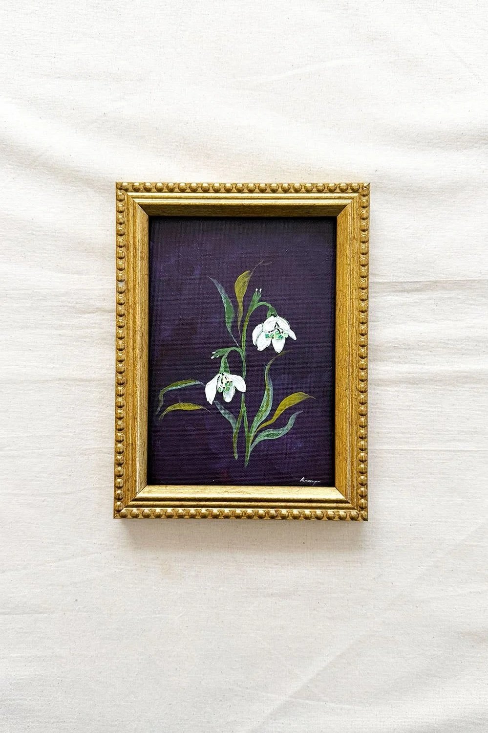 A framed painting of snowdrops on a dark purple background, housed in an ornate gold frame, displayed against a white fabric backdrop.