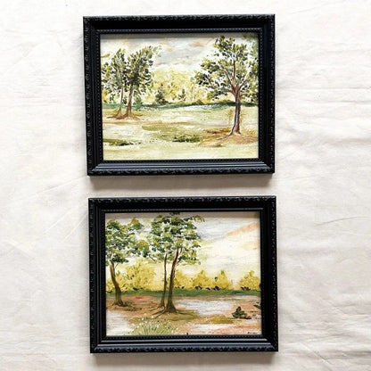 Two framed landscape original paintings of trees by a river, displayed vertically on a textured off-white fabric background. The frames are black with ornate detailing.
