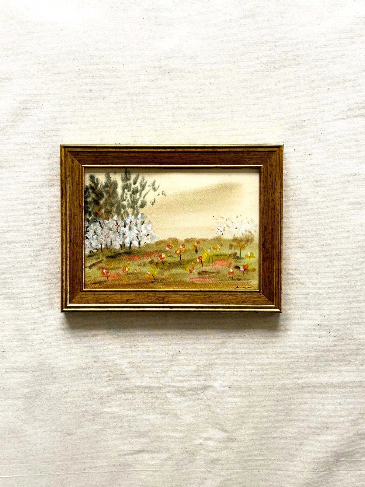 A small framed landscape painting on textured off-white paper. The painting depicts a grassy field with white flowering trees and a warm beige sky, enclosed in a brown wooden frame.