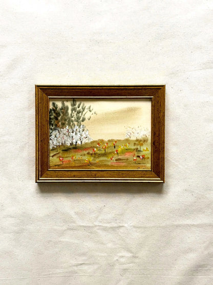 A small framed landscape painting on textured off-white paper. The painting depicts a grassy field with white flowering trees and a warm beige sky, enclosed in a brown wooden frame.