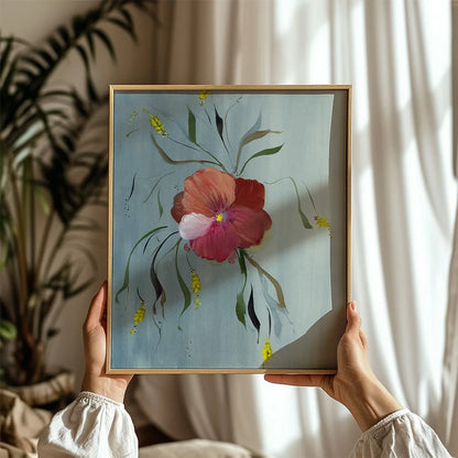 A person holds a square framed floral painting in a warm indoor setting. The artwork features a large pink and red flower with green leaves on a light blue background, enclosed in a thin gold frame.