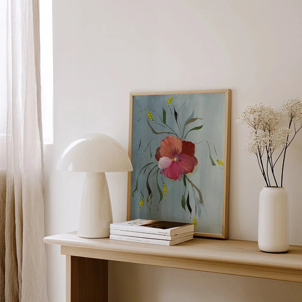 A framed botanical art print featuring a pink flower on a light blue background, styled on a light wood console table next to a white mushroom lamp and a white vase with dried flowers.
