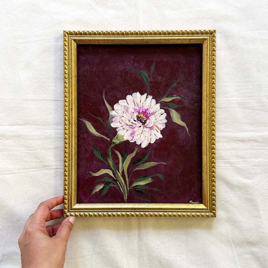 A framed vintage-style painting of a white and pink peony flower with green leaves on a deep burgundy background, housed in a decorative gold frame. Original by Ramya Rajiv