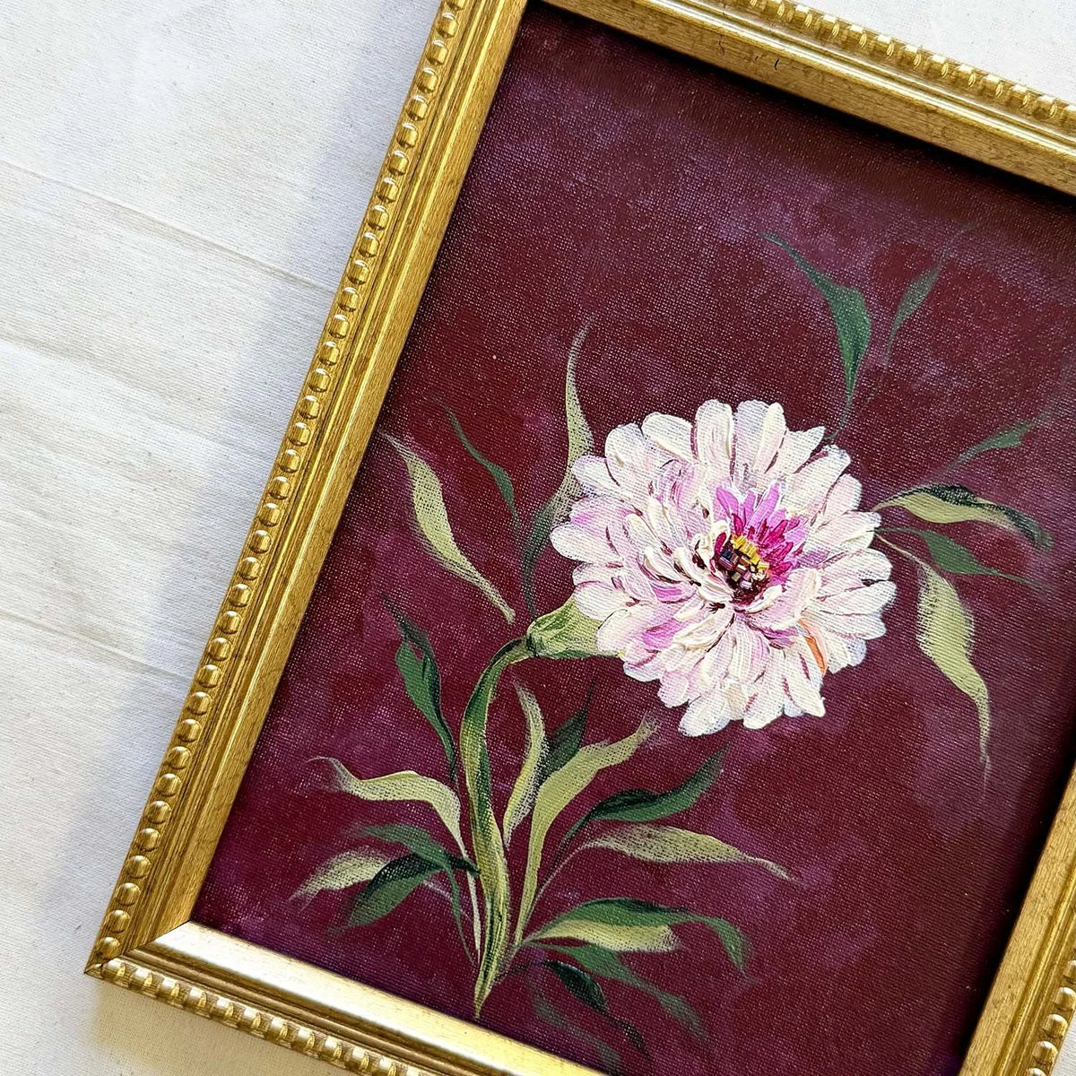 A framed vintage-style painting featuring a single white and pink peony flower with green leaves on a deep burgundy background, enclosed in an ornate gold frame.