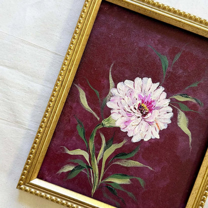 A framed vintage-style painting featuring a single white and pink peony flower with green leaves on a deep burgundy background, enclosed in an ornate gold frame.